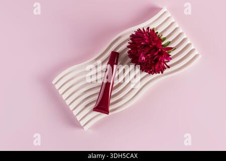 A vibrant red lip balm and a deep pink aster flower rest elegantly on a white, undulating textured tray against a soft pink background, creating a har Stock Photo