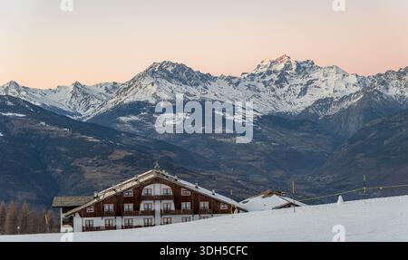 A picturesque winter scene in the Italian Alps, capturing a snow-covered chalet under soft pink skies surrounded by majestic snow-capped mountains. Tr Stock Photo