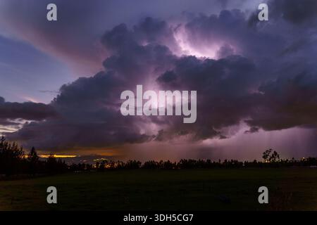 A dramatic thunderstorm at dusk with vibrant lightning illuminating dark clouds over a serene landscape. The contrasting colors highlight the power an Stock Photo