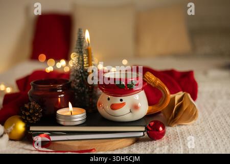 A cozy christmas, new year decor scene 6, featuring a snowman mug, candles, pine cone, red ornament, and fairy lights on a white knitted blanket with Stock Photo