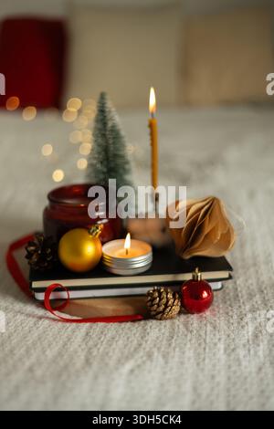 Festive scene with a lit candle, mini tree, and ornaments on books. The white and red accents create a cozy atmosphere on a soft, textured fabric back Stock Photo