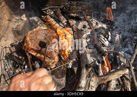 A steak grilling over an open flame, nestled among charred wood and coal, creating a rustic, smoky cooking atmosphere outdoors. The flames dance aroun Stock Photo