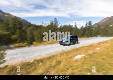 A dark SUV speeds along a gravel road in a mountainous area, surrounded by lush green pine trees and a clear blue sky, showcasing the thrill of off-ro Stock Photo