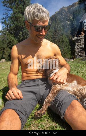 A man enjoys a sunny day in the mountains, sitting in the grass with his loyal dog resting on his lap. The serene outdoor setting offers a glimpse of Stock Photo