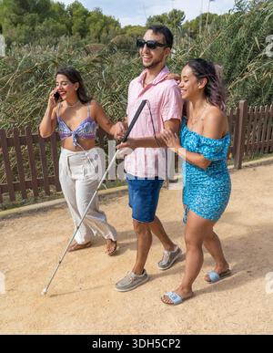 A group of three smiling friends, including a blind man with a cane, walking together happily along a park path on a sunny day, celebrating friendship Stock Photo