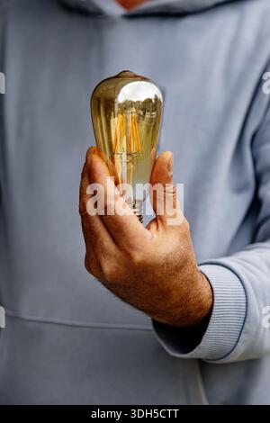 A man is holding a vintage style light bulb in his hand while wearing a light gray hoodie. The warm glow of the bulb contrasts with the soft fabric of Stock Photo