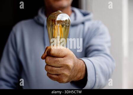 A close up of a man in a blue hoodie holding a vintage Edison style bulb in his hand, symbolizing creativity and innovation. The focus is on the bulb Stock Photo