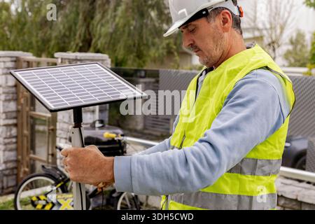 A man wearing a safety vest and helmet is holding a solar panel outdoors. The scene highlights renewable energy and sustainable practices in an open e Stock Photo