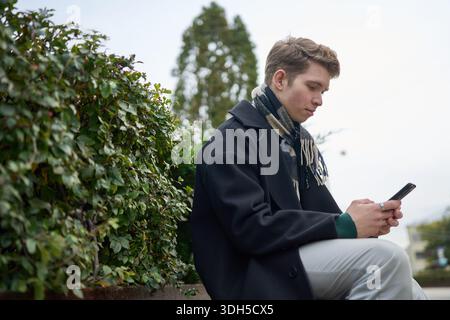 A young man sits outdoors in an urban setting during autumn, engrossed in his smartphone. He wears a dark coat and scarf, surrounded by greenery in a Stock Photo