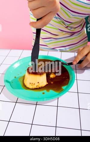A person uses a black plastic knife and fork to cut into a flan dessert on a vibrant blue plate. The background features a pink wall and a patterned t Stock Photo