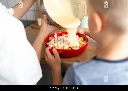 Close-up of two person preparing a homemade apple pie, pouring batter over sliced apples in a red dish. The cozy kitchen scene captures the joy of bak Stock Photo