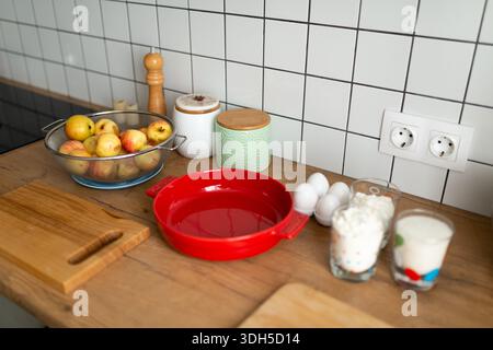 A cozy kitchen countertop is arranged with baking essentials including fresh apples in a colander, a red baking dish, eggs, flour, and sugar, creating Stock Photo