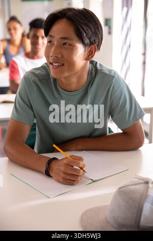 Asian teen writing with yellow pencil on spiral notebook at desk in class in light-green tee Stock Photo