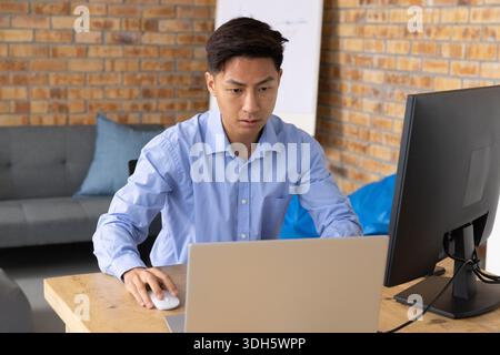 Asian man sitting at wooden desk in workspace, using laptop and external monitor Stock Photo