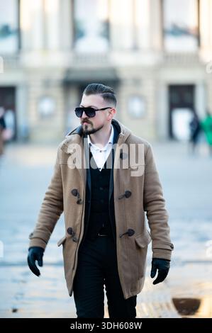 A trendy man walks confidently in a city square, wearing a warm coat and sunglasses, as people pass by in the vibrant afternoon light, enjoying the ur Stock Photo