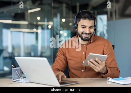 Young indian man wearing a headset and smiling, sitting at a desk and simultaneously using a laptop and a digital tablet, working in a modern office environment Stock Photo