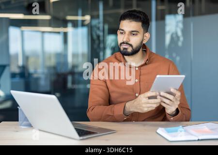 Young man concentrating on work, holding a digital tablet while sitting at a desk with a laptop, managing tasks in a modern corporate office environment Stock Photo