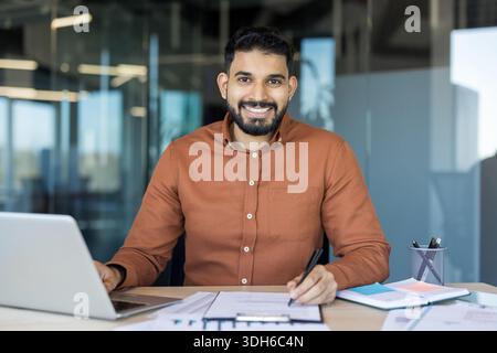 Young indian man with a beard smiling at the camera, working with a laptop and documents on his desk in a collaboration office space, representing success and professionalism Stock Photo