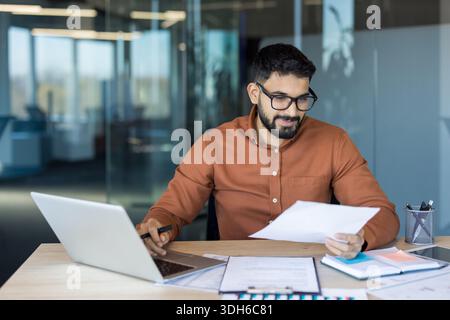 Young man in glasses and beard concentrating while working on a laptop and reviewing financial reports and paperwork, at a desk in a modern corporate office Stock Photo