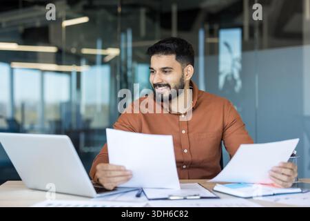 Young indian businessman at a modern office desk, smiling while reviewing reports and laptop, planning finances and strategy with confident, focused professionalism and productivity Stock Photo