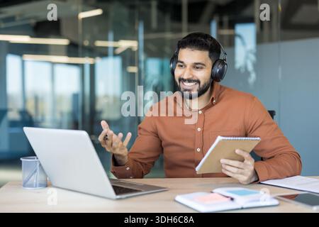 Young indian man wearing headphones and using laptop for a video call while working from a modern corporate office, actively discussing and taking notes from a notebook Stock Photo