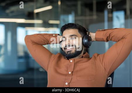 Happy bearded man in a casual shirt wearing headphones, smiling and leaning back in an office chair, enjoying a relaxing music break and calm, carefree moment at work Stock Photo