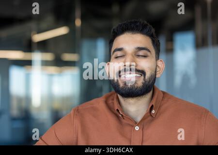 Young bearded entrepreneur finding peace and calm, closing eyes and smiling with happiness while practicing mindfulness at the modern corporate office to reduce stress and improve well-being Stock Photo