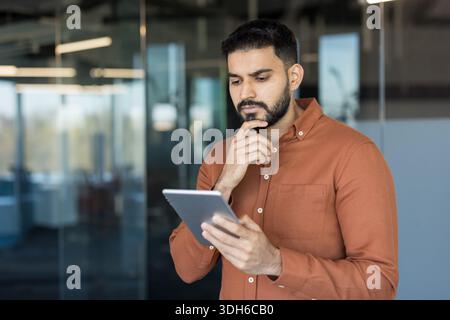 Young indian businessman looking down at a digital tablet, deep in thought while standing in a modern office, representing concepts of decision-making and focused business strategy Stock Photo