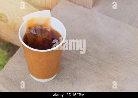 Black tea to-go. Paper cup with tea bag on a wooden stand. Breakfast takeaway. Top view. Stock Photo