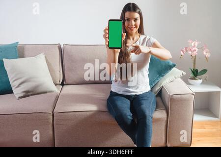 A woman with long hair sits on a couch showing a mobile device with a blank green screen. She smiles and points at the device in a living room with mo Stock Photo