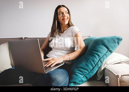 A woman sits on a couch with a laptop in front of her. She smiles while looking at the screen. Soft light comes through the window, creating a bright Stock Photo