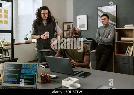 Young Black Man Smiling While Working on Laptop with Diverse Coworkers Stock Photo