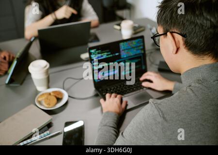 Young Asian Man Writing Code on Laptop While Collaborating with Colleagues Stock Photo