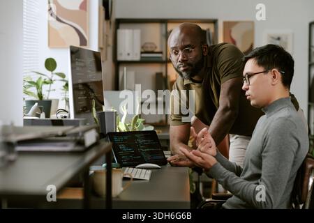 Young Black Man Supervising Young Adult Asian Man Coding on Laptop Stock Photo