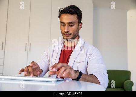 Asian man sitting at white desk typing on wireless keyboard in home office near storage cabinets. Concentration, productivity, modern, professional, cozy, minimalist, technology || Model released Stock Photo