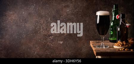 Glass of dark beer with foam head on dark wooden background with empty bottles and beer snacks Stock Photo