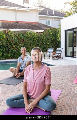 Senior couple practicing seated meditation on poolside patio with yoga mats and lounge chairs Stock Photo