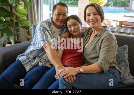Asian family sitting closely together on gray sofa in living room, holding hands near sliding door Stock Photo