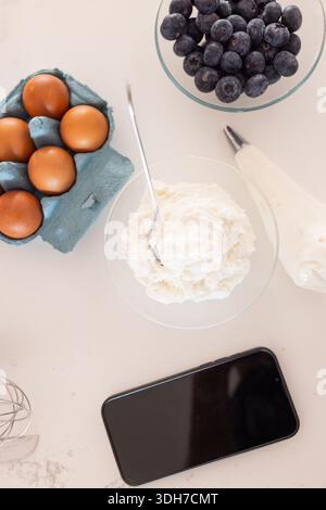 Glass bowl of whipped cream is sitting on light countertop, with egg carton, piping bag, smartphone Stock Photo