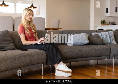 Woman in twenties wearing red T-shirt sitting on gray sofa working on silver laptop, copy space Stock Photo