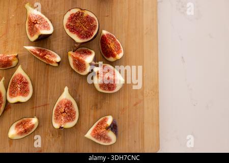 Halved and quartered figs are resting on wooden board by white stone countertop with juice stains Stock Photo