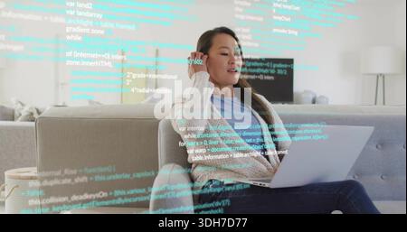 Coding Korean woman in cardigan at home on sofa, using silver laptop with cyan code overlay Stock Photo