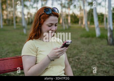 Smiling young red-haired woman sitting on a park bench using a smartphone outdoors. Stock Photo