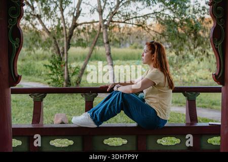 Young red-haired woman sitting sideways on a wooden gazebo bench in a park, relaxing and looking into the distance. Stock Photo