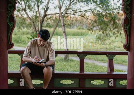 Young man writing in a notebook while sitting on a gazebo bench in a park, outdoor study and focus concept. Stock Photo