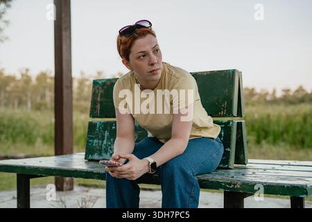 Young red-haired woman sitting on a green park bench holding a smartphone, looking away thoughtfully. Stock Photo