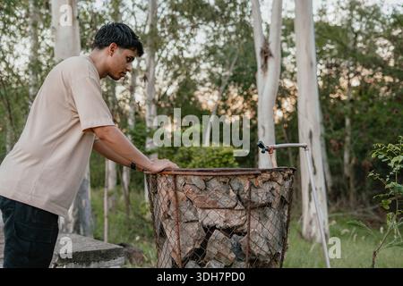 Young man reaching toward a wire firewood basket filled with logs in a park, practical outdoor task. Stock Photo