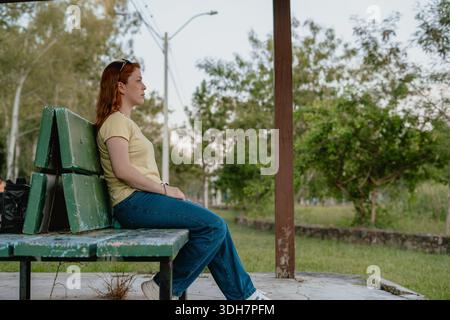 Young red-haired woman reclining on a green park bench in profile, relaxing in a quiet outdoor area. Stock Photo
