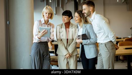 Four professionals gather in an office to review project details on a tablet. They share ideas and collaborate in a workspace with wooden tables and b Stock Photo