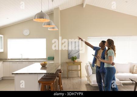 Diverse couple holding tablet pointing at ceiling and inspecting marble island at home, copy space Stock Photo